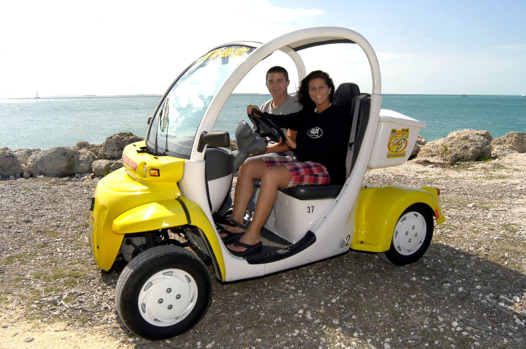 White and yellow two seater rental electric car with a woman and a young men aboard. They are parked in Key West by the Ocean
