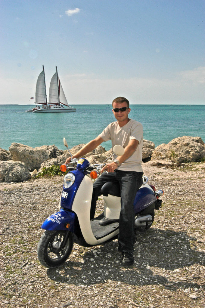 One guy on a one person blue and white scooter rental posing at Fort Zachary Taylor by the ocean with a sailboat in the background.
